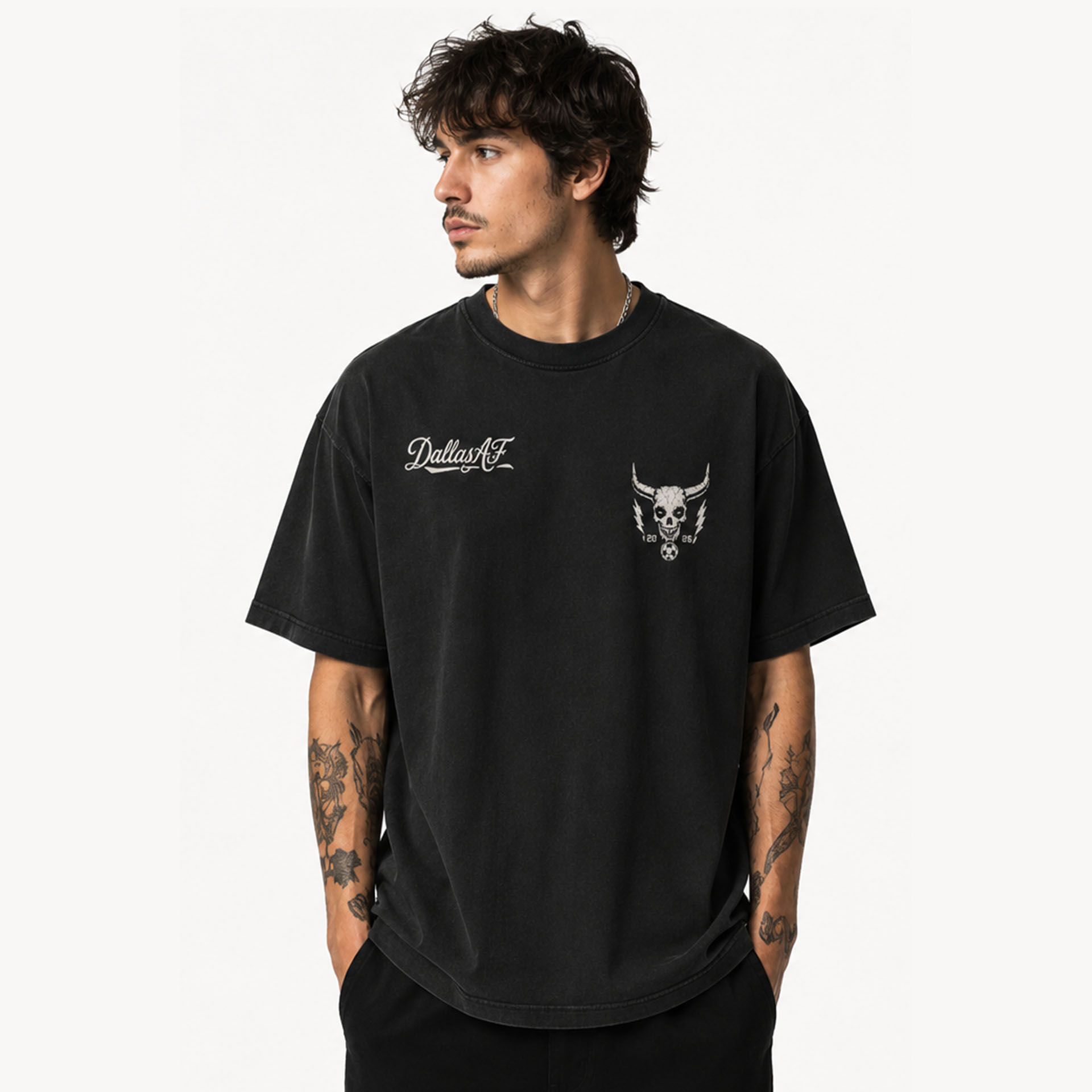 Man in a black oversized T-shirt facing left, with a Dallas logo and a skull graphic on the chest; tattoos visible on both forearms.